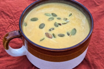 A brown and cream colored soup mug is filled with thick, velvety orange pumpkin soup, topped with salted pepitas atop a rust-colored tablecloth.