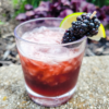 A dark-colored cocktail is served in a stout glass over ice, garnished on the rim with a lime wedge and three blackberries on a toothpick.