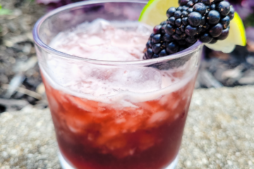 A dark-colored cocktail is served in a stout glass over ice, garnished on the rim with a lime wedge and three blackberries on a toothpick.
