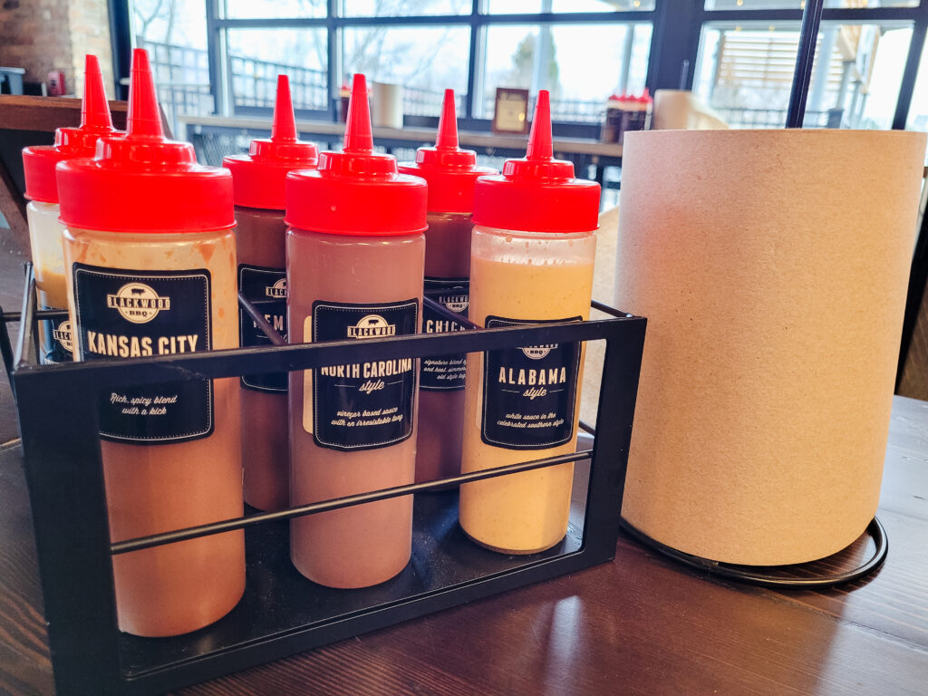 Six squeeze bottles of different regional Blackwood BBQ sauces are arranged in a simple black wrought iron basket on a table, next to a large roll of paper towels.
