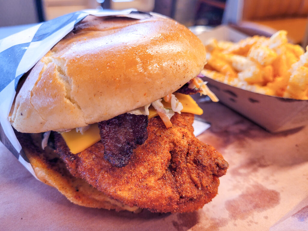 A hot chicken sandwich with cheese and coleslaw sits on a paper-lined tray, with golden french fries just behind it.