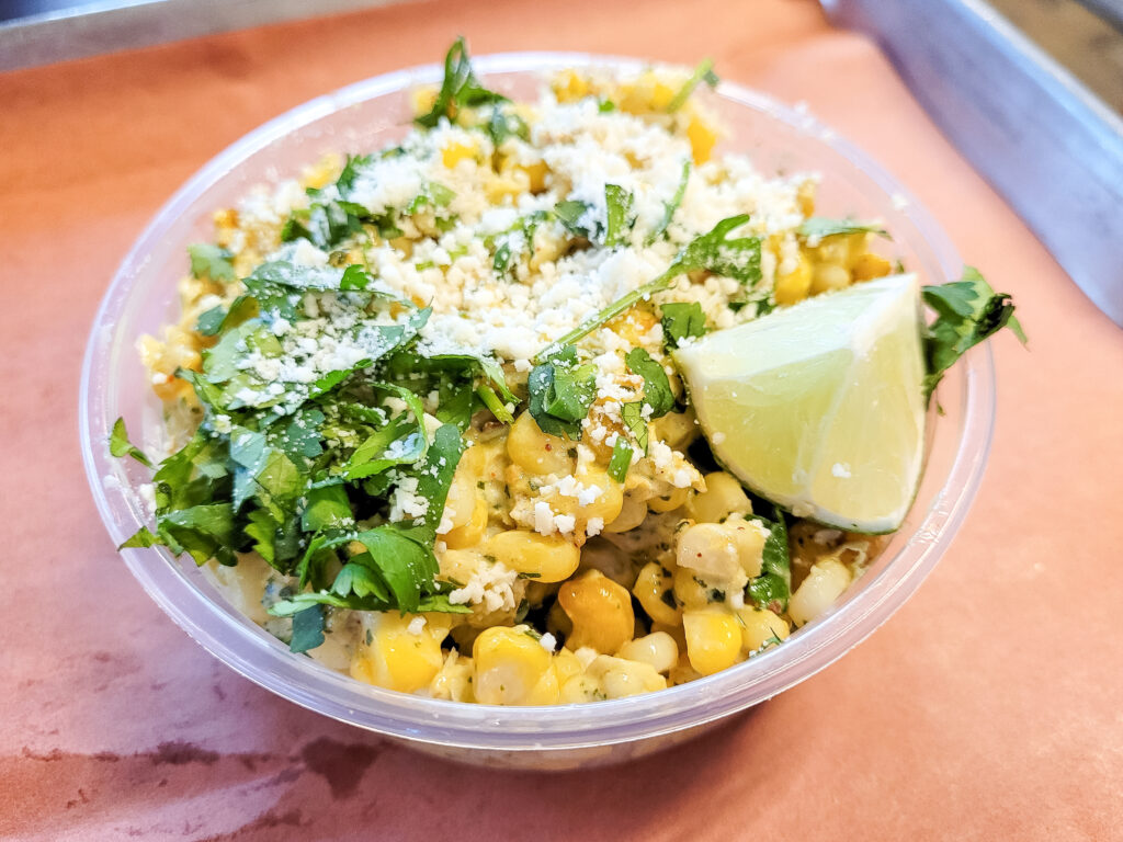 An individual-sized plastic dish of corn with a topping of chopped cilantro and crumbled cotija cheese, garnished with a lime wedge.