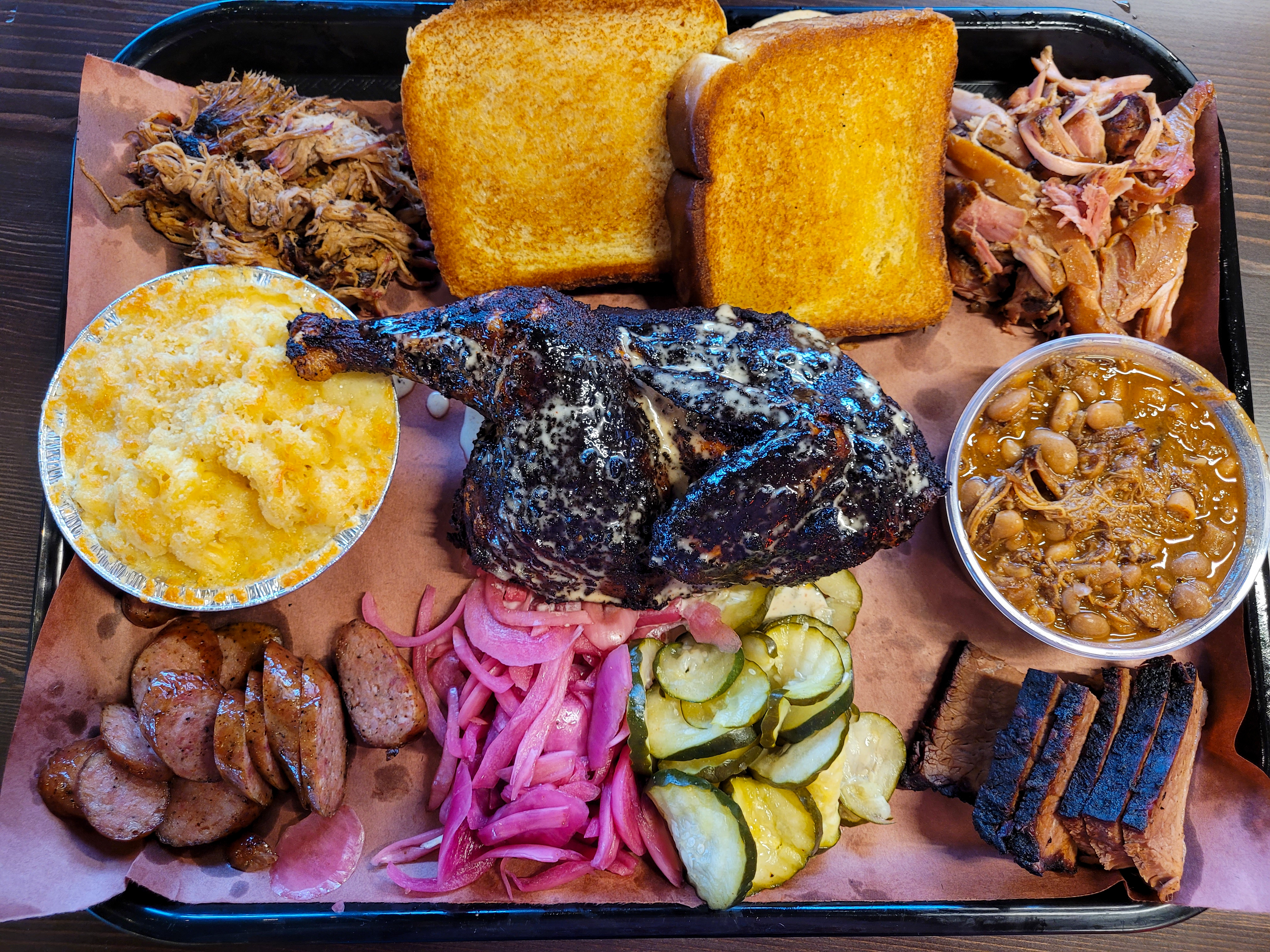 A paper-lined tray covered with barbequed meats and sides, including pulled pork, pulled chicken, brisket, sliced sausage, half a blackened chicken, macaroni and cheese, pork and beans, pickled onions, pickles, 2 slices of Texas toast, and two corn muffins.