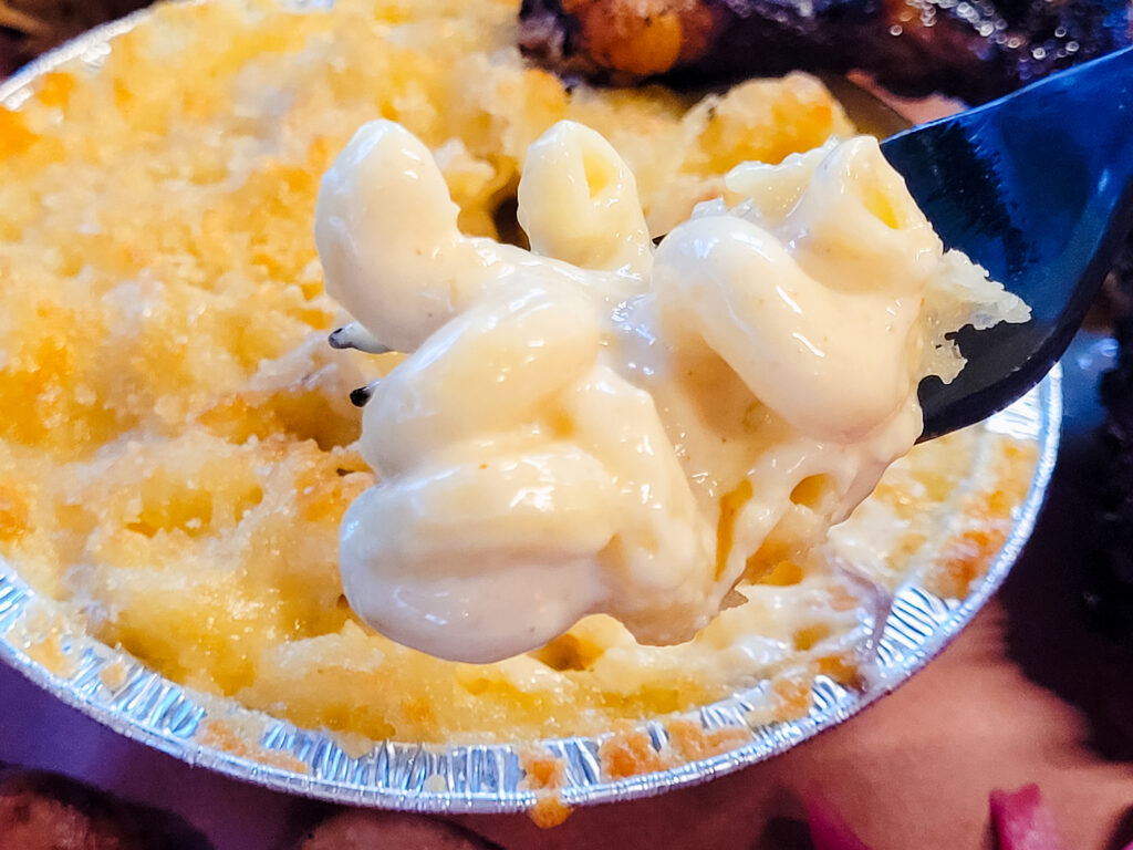 A forkful of creamy, pale yellow macaroni and cheese graces the foreground while a small circular tin of macaroni and cheese crusted with panko can be seen in the background.