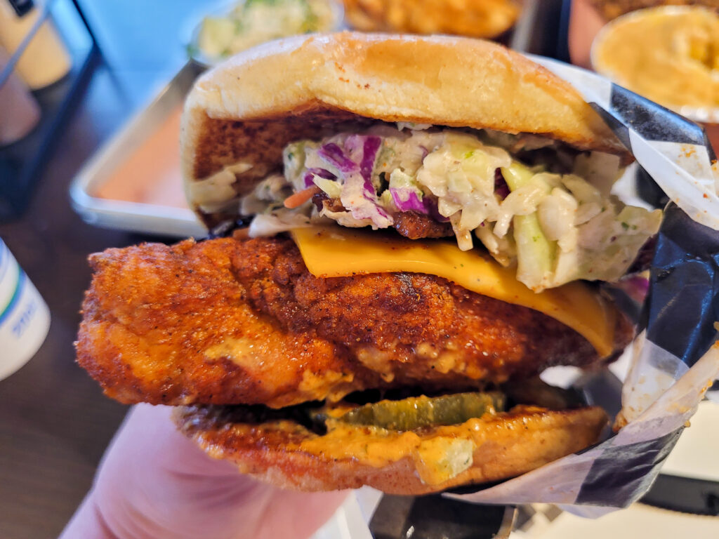 One hand holds a hot chicken sandwich, consisting of a large spicy breaded chicken breast on a Kaiser roll, topped with a slice of American cheese and coleslow.
