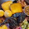 The Sampler Platter is shown, consisting of chicken, pulled pork, brisket, corn muffins, Texas toast, pork and beans, mac and cheese, and pickles.