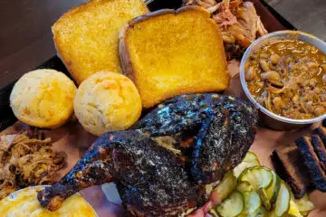 The Sampler Platter is shown, consisting of chicken, pulled pork, brisket, corn muffins, Texas toast, pork and beans, mac and cheese, and pickles.