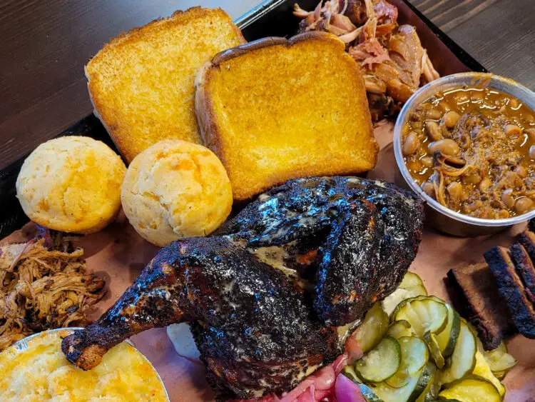 The Sampler Platter is shown, consisting of chicken, pulled pork, brisket, corn muffins, Texas toast, pork and beans, mac and cheese, and pickles.