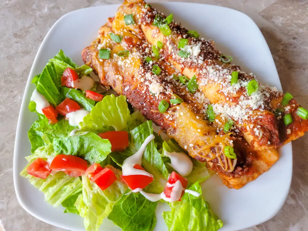 A square white dinner plate is half-filled with a bright green lettuce and tomato salad, drizzled with white dressing, and two cheese enchiladas laying side-by-side and topped with sauce, sliced green onions, and crumbled cotija cheese.