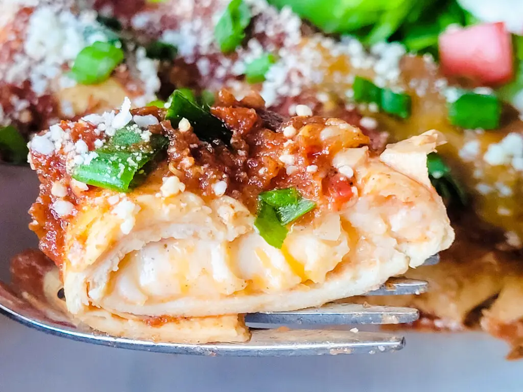 A slice of enchilada on a fork, the center oozing with melted cheese and topped with sauce, green onion slices, and crumbled cotija cheese.