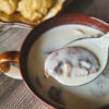 A dark brown mug of velvety white soup takes up most of the screen, with a spoon of soup and a single mushroom hovers over the soup.