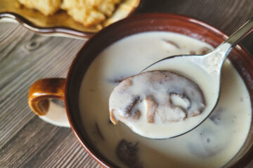 A dark brown mug of velvety white soup takes up most of the screen, with a spoon of soup and a single mushroom hovers over the soup.