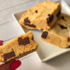 Two square-cut golden bar cookies with chocolate chunks lie on a square white plate, on slightly stacked over the other, while a lady's hand with long, red nails hold a third cookie square to the camera, showcasing a side view of its chocolate chunks.