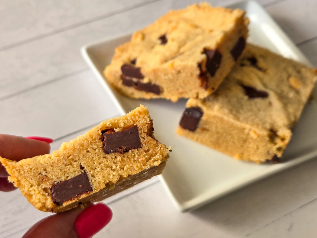 Two square-cut golden bar cookies with chocolate chunks lie on a square white plate, on slightly stacked over the other, while a lady's hand with long, red nails hold a third cookie square to the camera, showcasing a side view of its chocolate chunks.