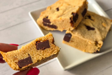 Two square-cut golden bar cookies with chocolate chunks lie on a square white plate, on slightly stacked over the other, while a lady's hand with long, red nails hold a third cookie square to the camera, showcasing a side view of its chocolate chunks.