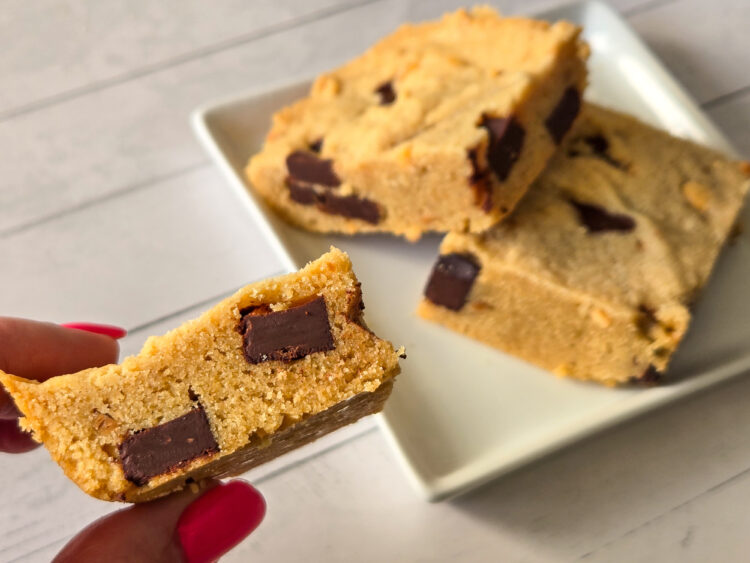 Two square-cut golden bar cookies with chocolate chunks lie on a square white plate, on slightly stacked over the other, while a lady's hand with long, red nails hold a third cookie square to the camera, showcasing a side view of its chocolate chunks.