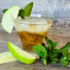 A cocktail glass is filled with a golden liquid and ice cubes, with a green apple slice and three mint leaves garnishing the glass, while more mint leaves and apple slices lay at the base of the glass.