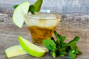 A cocktail glass is filled with a golden liquid and ice cubes, with a green apple slice and three mint leaves garnishing the glass, while more mint leaves and apple slices lay at the base of the glass.