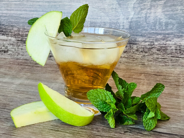 A cocktail glass is filled with a golden liquid and ice cubes, with a green apple slice and three mint leaves garnishing the glass, while more mint leaves and apple slices lay at the base of the glass.