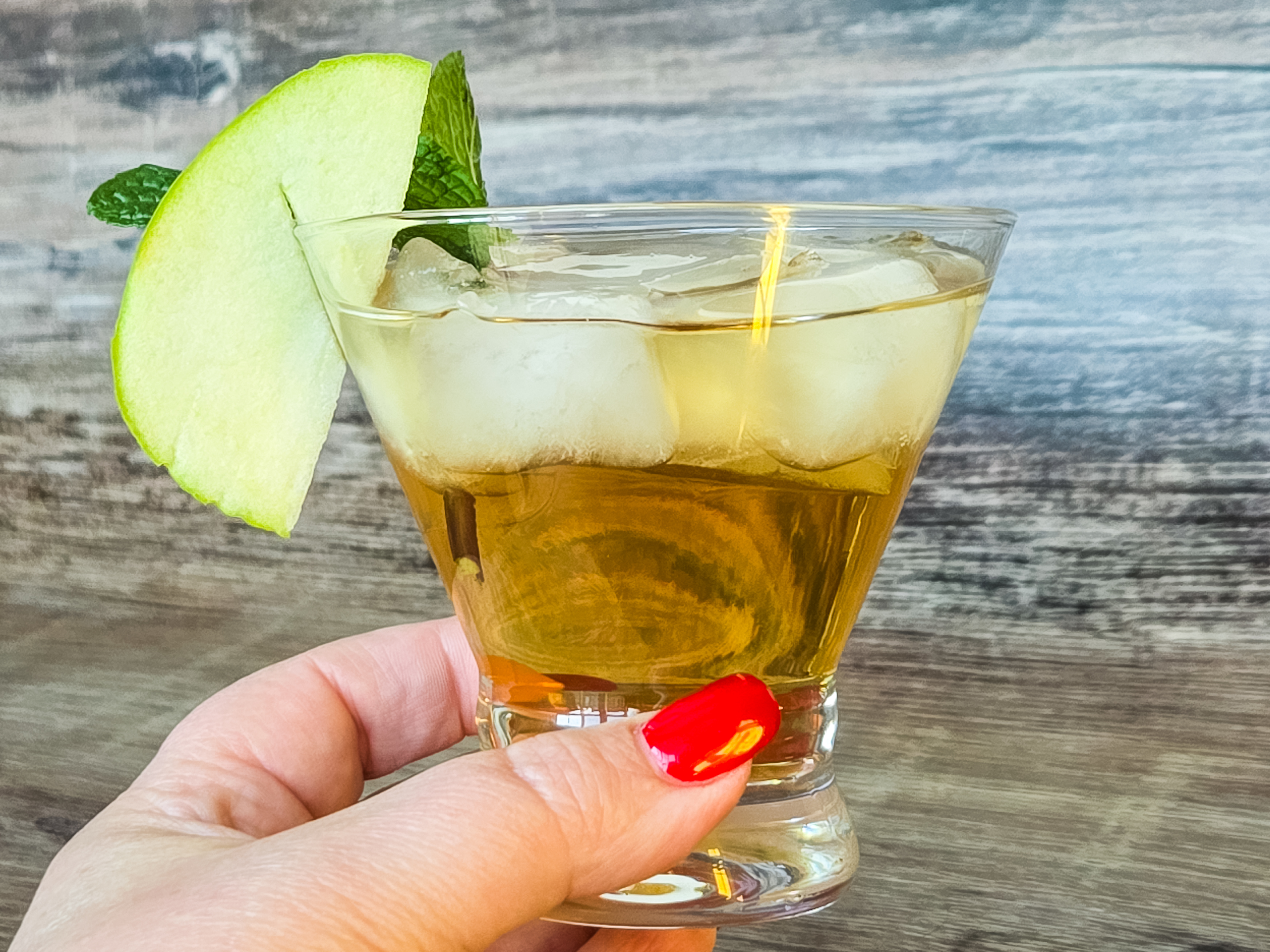 A woman's hand with red fingernails holds a cocktail glass of golden liquid over ice in front of a wooden panel.