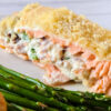 A slice of pink salmon, stuffed with a white, creamy mixture of cream cheese, bacon, and jalapeno pepper and topped with panko, lies horizontally on a white plate next to fresh asparagus.