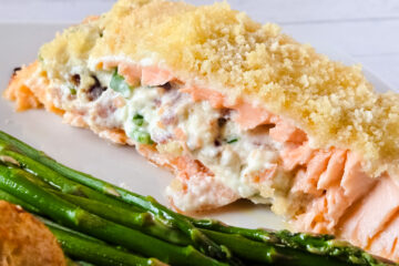 A slice of pink salmon, stuffed with a white, creamy mixture of cream cheese, bacon, and jalapeno pepper and topped with panko, lies horizontally on a white plate next to fresh asparagus.