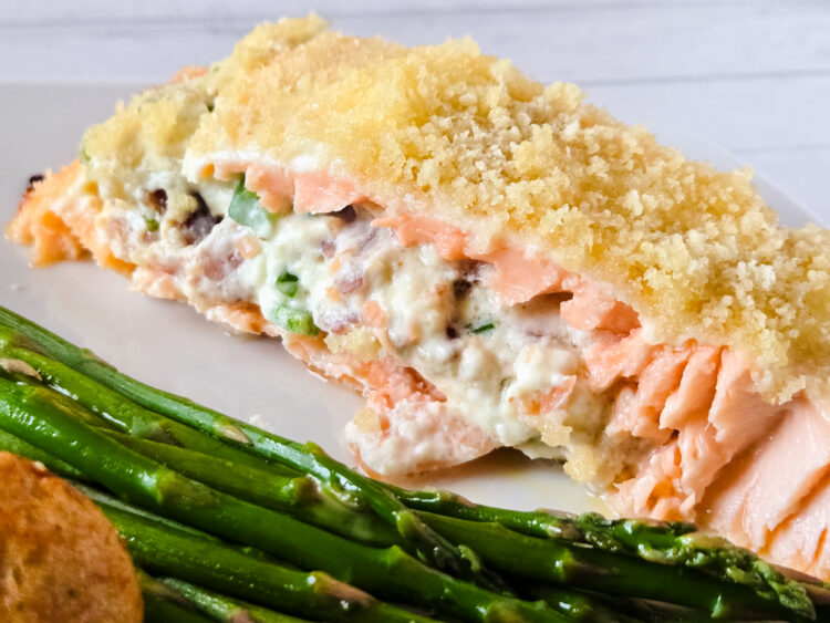 A slice of pink salmon, stuffed with a white, creamy mixture of cream cheese, bacon, and jalapeno pepper and topped with panko, lies horizontally on a white plate next to fresh asparagus.